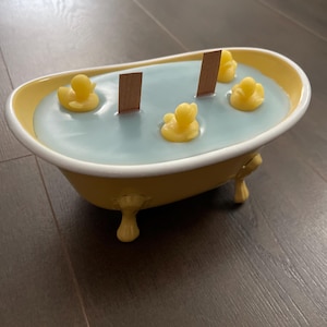 Puede incluir: Una vela amarilla con forma de bañera con un interior de cera azul claro. La vela presenta tres patitos de goma amarillos y dos mechas de madera. La bañera tiene patas decorativas y un borde blanco.
