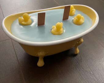 May include: A yellow bathtub-shaped candle with a light blue wax interior. The candle features three yellow rubber duckies and two wooden wicks. The bathtub has decorative feet and a white rim.