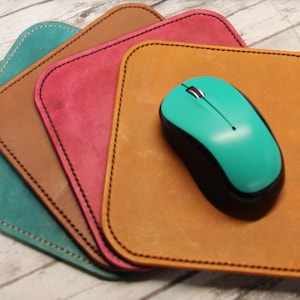 May include: A teal wireless mouse sits on a tan leather mousepad with black stitching. Three other mousepads in teal, brown, and pink are stacked behind it. The mousepads have rounded corners.