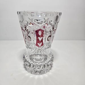 May include: A clear crystal vase with a cut glass design. The vase has a flared top and a rounded base. Red panels with white floral designs are visible through the clear glass. The vase is approximately 6 inches tall.
