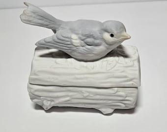 May include: A ceramic trinket box with a light gray bird perched on top. The bird has detailed feather markings and a small yellow beak. The box is white and shaped like a log, with a hinged lid. A decorative item.