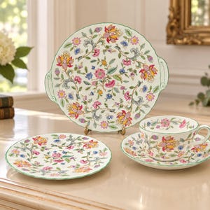 May include: A ceramic tea set featuring a cake plate, a small plate, and a teacup with a saucer. Each piece is white with a colourful floral pattern in shades of pink, yellow, blue, and green, finished with a scalloped green rim detail.