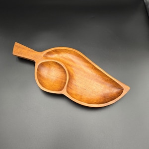 May include: A wooden leaf-shaped serving tray with a circular indentation and a larger, elongated section. The tray is made of a light brown wood and has a smooth, polished finish. The tray is designed for serving snacks or small dishes.