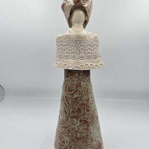 May include: A ceramic sculpture of a woman with a head covering and a lace-trimmed shawl. The figure has a cream-colored face and shoulders. The dress is a reddish-brown color with a floral pattern and a light green overlay.