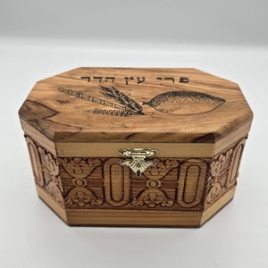 May include: An octagonal wooden box with a natural grain finish. The lid features a laser-engraved etrog and lulav illustration with Hebrew text. The sides are decorated with intricate carved floral patterns and a silver-toned metal latch closure.