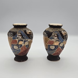May include: A pair of small, black ceramic vases featuring hand-painted figures in traditional attire. The designs incorporate gold, blue, and red accents with intricate patterns. Each vase has a narrow neck and a flared rim with gold trim.