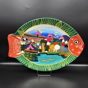 Op de afbeelding: Een handbeschilderde, keramische visvormige schaal met een levendige scène. De schaal heeft een groene rand, een oranje staart en oog, en een centraal landschap met figuren, dieren en een waterpartij. Het kunstwerk is kleurrijk en gedetailleerd.
