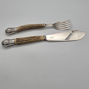 May include: A silver fork and a wide silver knife with textured, light brown antler handles. Both utensils feature ornate, polished silver caps at the ends of the handles. The knife blade has a visible inscription near the handle base.
