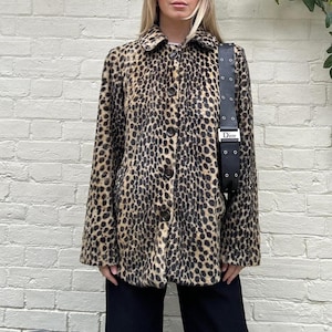 May include: A long-sleeved jacket featuring a brown and black leopard print pattern, a classic collar, and a front button closure. A black shoulder bag with a silver-toned buckle strap and the brand name Dior printed on the hardware is worn over one shoulder.