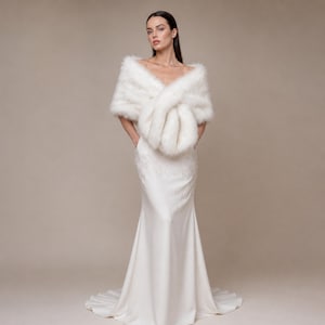 May include: A floor-length white gown featuring a smooth, solid fabric skirt and a textured, white faux fur wrap draped over the shoulders. The wrap is crossed at the front, creating a voluminous, soft appearance that contrasts with the sleek, fitted bodice.