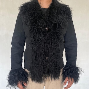 May include: A black jacket featuring a shaggy fur collar and cuffs. The jacket has a button closure and a tailored fit. The sleeves are long, and the cuffs are trimmed with the same fur as the collar. The jacket is worn with beige trousers.