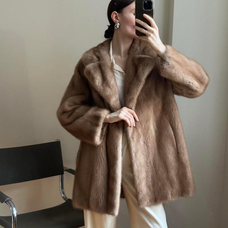 May include: A light brown fur coat with a classic collar and long sleeves. The coat is knee-length and has a soft, textured appearance. The coat is worn over a white shirt and cream-colored pants.
