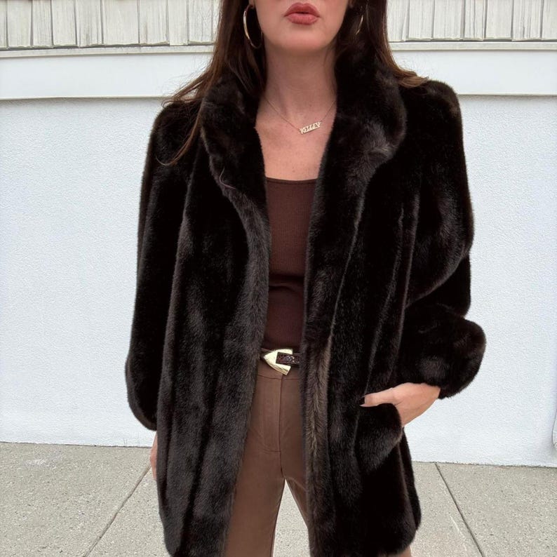 May include: A dark brown faux fur coat with a wide collar and long sleeves. The coat has a classic design and appears to be made of a soft, plush material. The coat is worn over a brown top and pants.