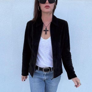 Könnte beinhalten: Schwarze Wildlederjacke mit Reverskragen und Reißverschluss. Die Jacke wird über einem weißen Tanktop und einer blauen Jeans getragen. Ein schwarzer Gürtel mit silberner Schnalle und eine Kreuz-Anhänger-Halskette vervollständigen den Look.
