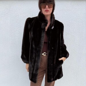 May include: A dark brown faux fur coat with a high collar and button closure. The coat has long sleeves and a mid-length cut. The coat is a solid color and appears to be made of a soft, plush material. The coat has pockets.