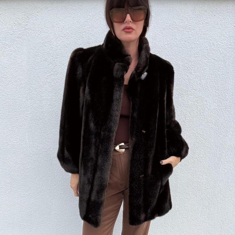 May include: A dark brown faux fur coat with a high collar and button closure. The coat has long sleeves and a mid-length cut. The coat is a solid color and appears to be made of a soft, plush material. The coat has pockets.