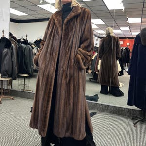 May include: A full-length brown fur coat with a wide collar and long sleeves. The coat is open in the front, revealing a black outfit underneath. The coat has a luxurious, textured appearance and is displayed in a clothing store.