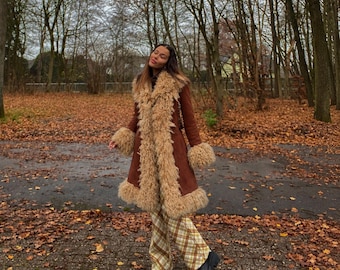 May include: A brown suede coat featuring long, shaggy beige faux fur trim along the lapels, cuffs, and hemline. The coat is paired with beige and brown plaid trousers and black platform shoes, creating a layered look for cooler weather.