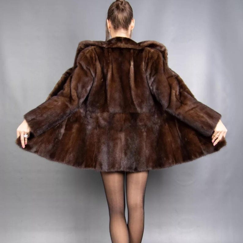 May include: A dark brown fur coat with a wide collar and flared hem. The coat is a rich, dark brown colour and appears to be made of soft fur. The coat is open, revealing the lining. The person wearing the coat is facing away from the camera.