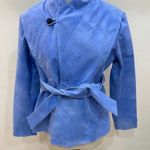 May include: A light blue suede jacket with a wrap-around design and a matching belt. The jacket features a high collar and a single black button closure. The sleeves are long, and the jacket is displayed on a mannequin.