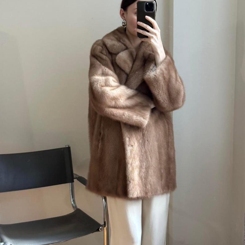 May include: A brown fur coat with a wide collar and long sleeves. The coat is knee-length and appears to be made of a soft, plush material. The coat is buttoned up and worn over white pants.