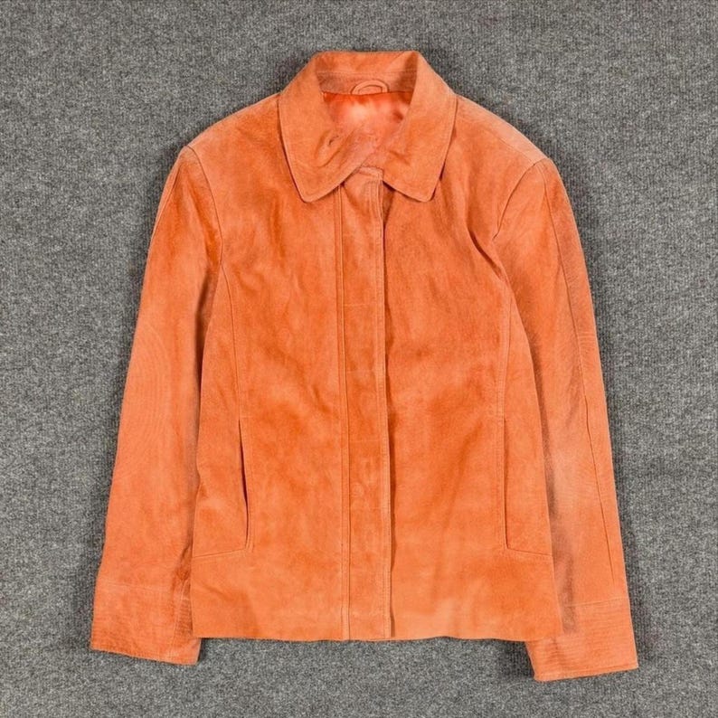 Peut inclure: Une veste en daim orange vif dot&eacute;e d'un col pointu classique, de manches longues et d'une patte de boutonnage avant dissimul&eacute;e. Le v&ecirc;tement pr&eacute;sente une coupe structur&eacute;e avec des coutures verticales sur les panneaux avant et deux poches.