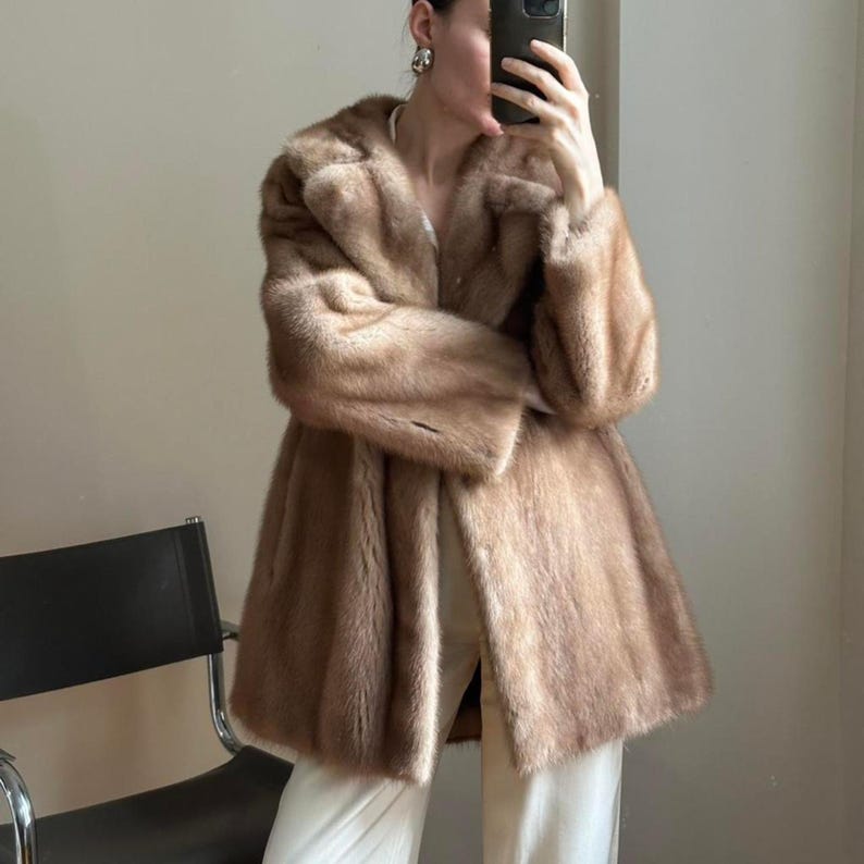 May include: A brown fur coat with a wide collar and long sleeves. The coat is knee-length and has a flared silhouette. The fur appears soft and luxurious, with a light brown color. The coat is open in the front, revealing white pants.