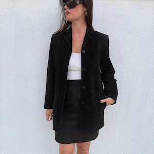 Könnte beinhalten: Ein schwarzer Wildlederblazer mit drei Knöpfen, kombiniert mit einem passenden schwarzen Wildlederrock. Das Outfit wird über einem weißen Oberteil getragen. Der Blazer hat ein klassisches, maßgeschneidertes Design. Der Gesamteindruck ist schick und anspruchsvoll.