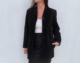 May include: A black suede blazer with three buttons, paired with a matching black suede skirt. The outfit is worn over a white top. The blazer has a classic, tailored design. The overall look is chic and sophisticated.