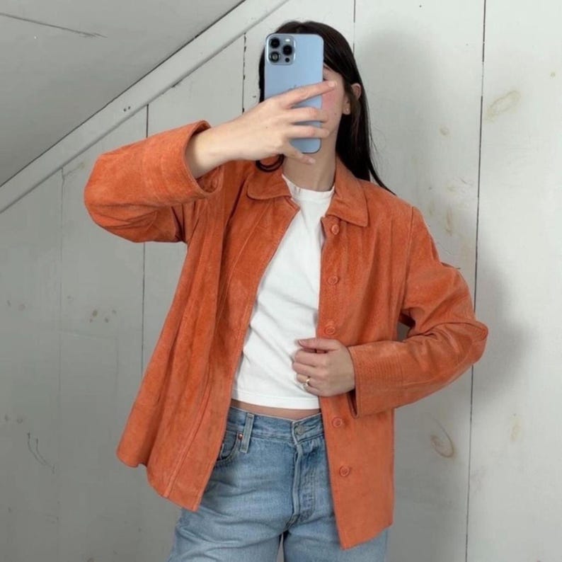 Peut inclure: Une veste en daim orange vif dot&eacute;e d'un col classique, de manches longues et d'une fermeture &agrave; boutons sur le devant. La veste est port&eacute;e sur un t-shirt blanc uni et un jean bleu clair. Le v&ecirc;tement pr&eacute;sente une finition textur&eacute;e et un ourlet droit.