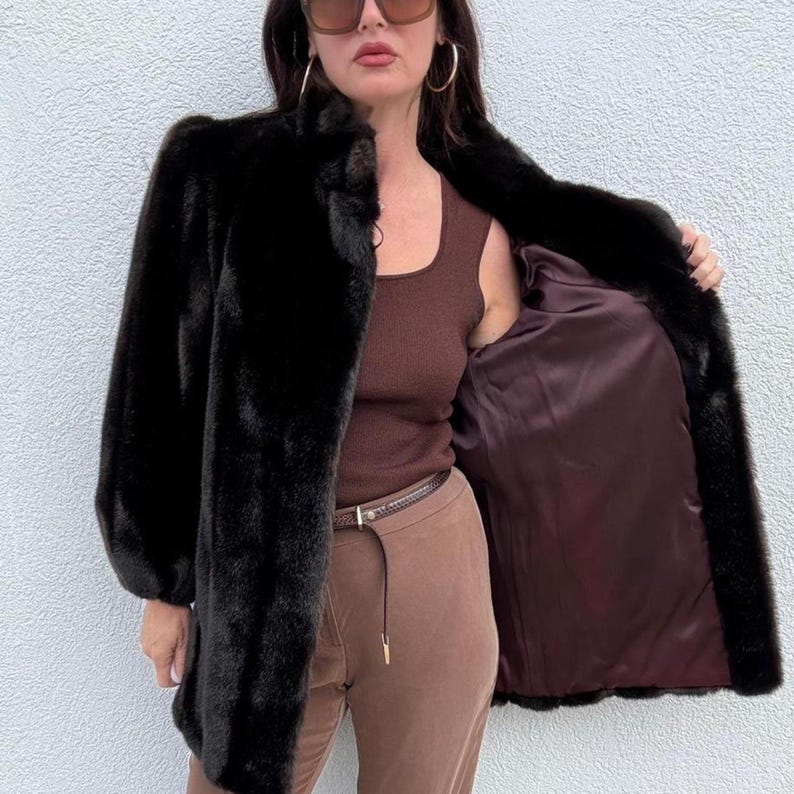 May include: A black faux fur coat with a high collar and long sleeves. The coat is open, revealing a dark brown satin lining. The person is wearing brown pants and a brown top. The coat is a classic outerwear piece.