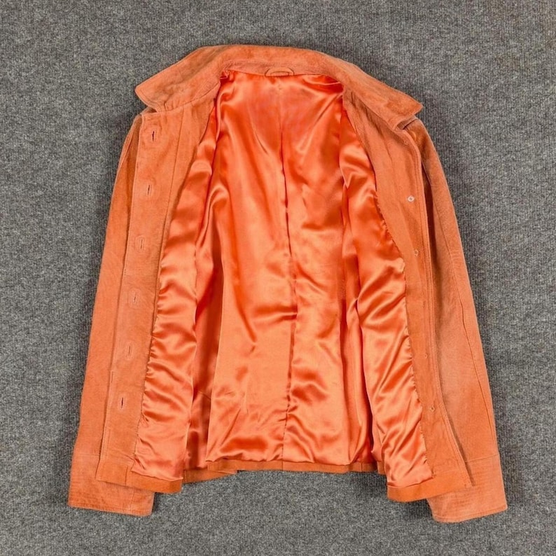 Peut inclure: Une veste en daim orange dot&eacute;e d'un col classique, de manches longues et d'une fermeture boutonn&eacute;e sur le devant. L'int&eacute;rieur est enti&egrave;rement doubl&eacute; d'un tissu satin&eacute; orange assorti, offrant une finition lisse &agrave; la structure du v&ecirc;tement.