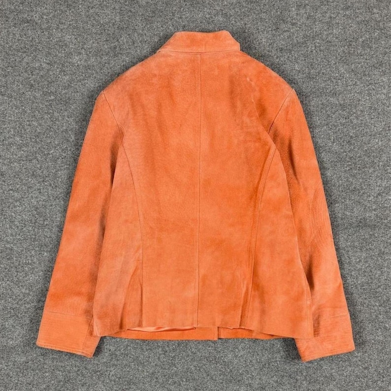 Peut inclure: Une veste en daim orange uni dot&eacute;e d'un col structur&eacute;, de manches longues avec poignets et d'une couture verticale centrale dans le dos. Le v&ecirc;tement pr&eacute;sente une texture mate lisse et une coupe ajust&eacute;e avec des panneaux visibles sur l'ensemble.