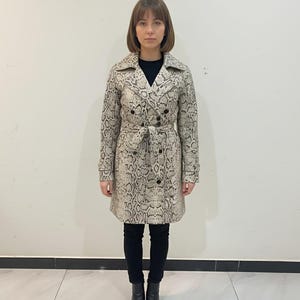 May include: A snakeskin-patterned trench coat in beige, grey, and black. The double-breasted coat has a belt, a notched collar, and black buttons. The model is wearing black trousers and boots. The coat's length is approximately 100 cm.