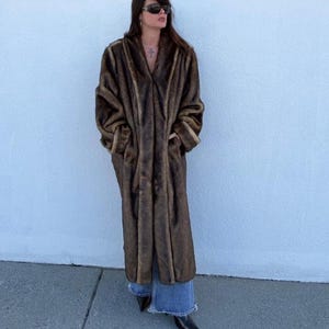 May include: A long, brown faux fur coat with vertical stripes. The coat has a V-neck and pockets. The coat is worn over blue denim jeans and black boots. The coat is a vintage style.