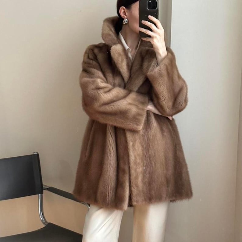 May include: A brown faux fur coat with a wide collar and long sleeves. The coat is knee-length and has a flared silhouette. The coat is a warm, neutral color and appears to be made of a soft, plush material. The coat is a stylish outerwear option.