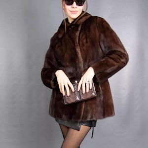 May include: A dark brown fur coat with a classic collar and button closure. The coat is paired with a small, brown, textured clutch. The overall look is completed with sunglasses and a black skirt.