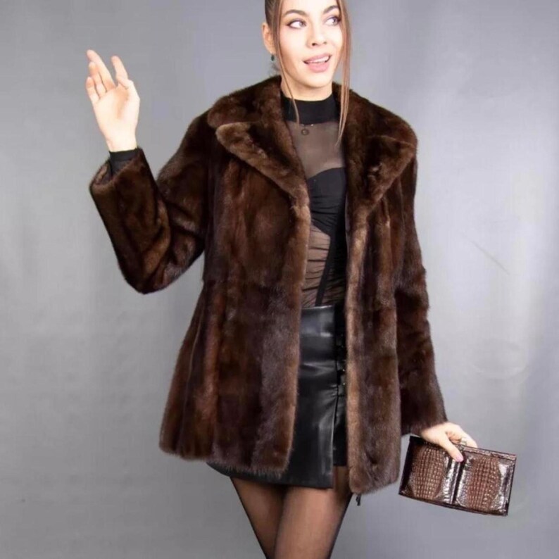 May include: A brown fur coat with a wide collar and long sleeves. The coat is knee-length and has a slightly flared shape. A brown leather clutch is held in the hand.