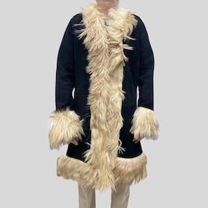 Può includere: Un cappotto in camoscio nero con finiture in finta pelliccia color crema. Il cappotto presenta finiture in pelliccia intorno al colletto, sul davanti e sui polsini e sull'orlo. Il cappotto è lungo fino al ginocchio e ha un'estetica vintage.