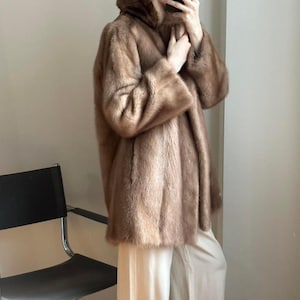 May include: A brown fur coat with a hood. The coat has long sleeves and a soft, textured appearance. The coat is knee-length and appears to be made of a luxurious material. The model is wearing light-colored pants.