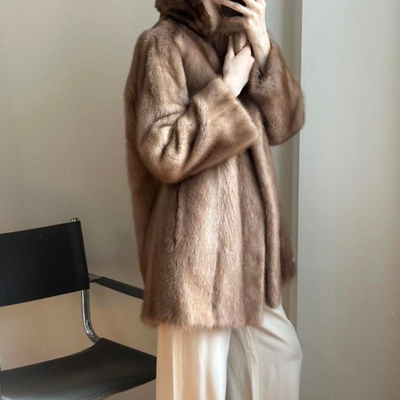 May include: A brown fur coat with a hood. The coat has long sleeves and a soft, textured appearance. The coat is knee-length and appears to be made of a luxurious material. The model is wearing light-colored pants.