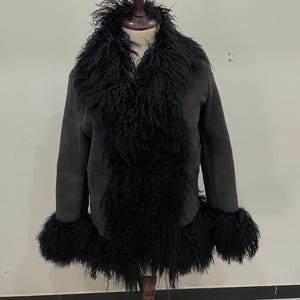 May include: A dark grey jacket with a black, shaggy fur collar and cuffs. The jacket is displayed on a wooden mannequin. The jacket has a suede-like texture and a V-neck design. The fur trim adds a luxurious touch to the outerwear.