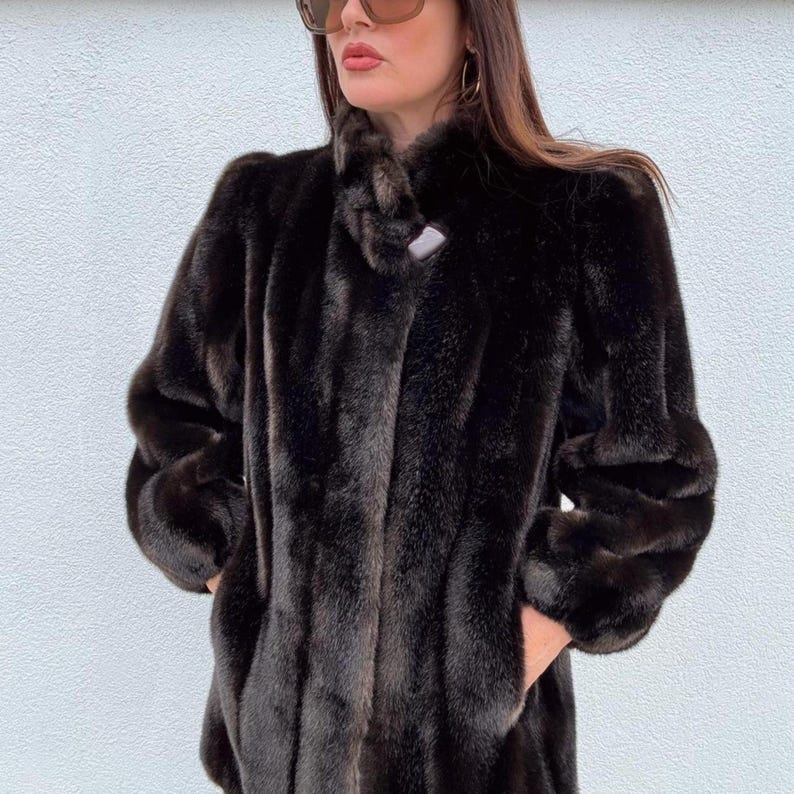 May include: A dark brown faux fur coat with a high collar and a single button closure. The coat has long sleeves and pockets. The coat is worn by a person. The background is a plain white wall.