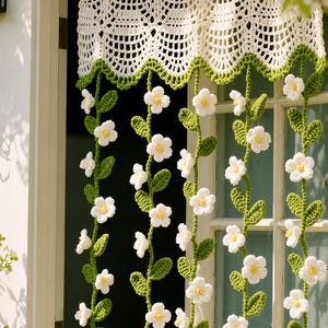 Puede incluir: Una cenefa de ganchillo color crema con borde festoneado y ribete verde, que presenta múltiples hilos verticales colgantes de enredaderas verdes tejidas con flores blancas de centros amarillos, suspendida sobre una puerta.