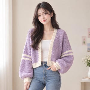 Könnte beinhalten: Eine lavendelfarbene Häkelstrickjacke mit langen, voluminösen Ärmeln, die jeweils zwei horizontale cremefarbene Streifen aufweisen. Das Kleidungsstück hat einen cremefarbenen Rippstricksaum entlang der Vorderöffnung, an den Ärmelbündchen und am Saum.