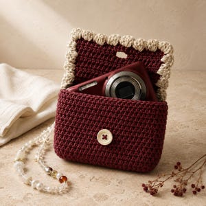 May include: A dark red crocheted pouch with a cream-coloured scalloped edge on the flap and a single cream button closure. A dark red digital camera is tucked inside the pouch, and a long beaded strap with clear and amber beads is attached.