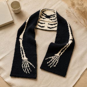 Könnte beinhalten: Ein schwarzer Strickschal mit einem weißen Intarsienmuster, das einen menschlichen Brustkorb in der Mitte sowie skelettierte Arm- und Handknochen zeigt, die sich an beiden Seiten nach unten erstrecken. Das Design ist kontrastreich und detailliert.