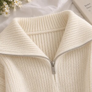 May include: A cream-coloured knit jumper featuring a wide fold-over collar and a silver-toned metal zip that extends from the neckline down the centre. The garment is constructed with a vertical ribbed texture throughout the fabric.