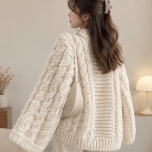 Könnte beinhalten: Ein cremefarbener Zopfmuster-Pullover mit hohem Kragen, langen Ärmeln und geripptem Saum. Der Pullover zeigt ein markantes vertikales Zopfmuster an den Ärmeln und Seiten, kombiniert mit einer glatten beigen Umhängetasche mit Riemen.
