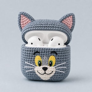 May include: A gray crocheted protective case for wireless earbuds, designed to resemble a cat face with pointed ears featuring pink inner details, yellow eyes, a white muzzle, a black nose, and white embroidered whiskers on the front.